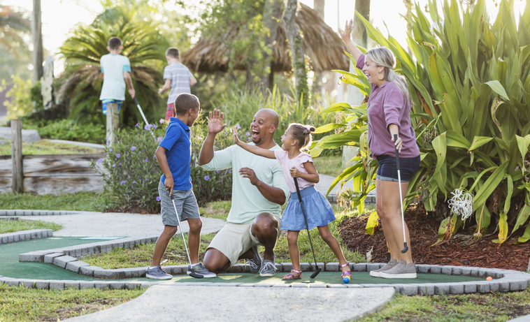 An interracial family having fun together playing miniature golf. The African-American father and Caucasian mother are in their 30s. The children are 9 and 5 years old. Dad and sister are giving the boy high fives for his winning putt.